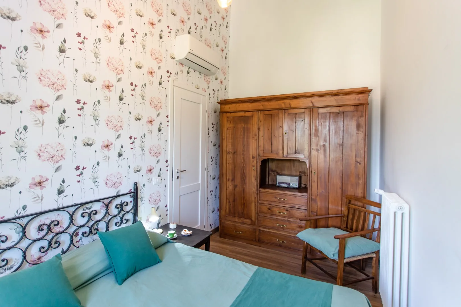 A bedroom with a bed, floral wallpaper, and a large wooden wardrobe.