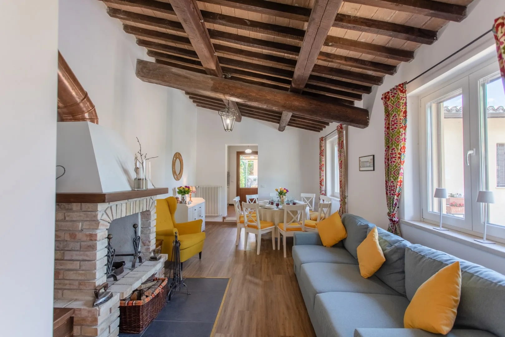 A bright living and dining room with a fireplace and exposed wooden ceiling beams.
