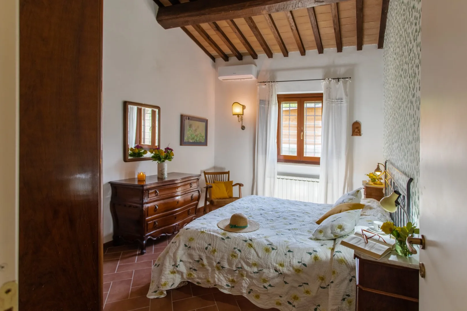 A bedroom with a bed beneath a wooden-beamed ceiling and a window with curtains.