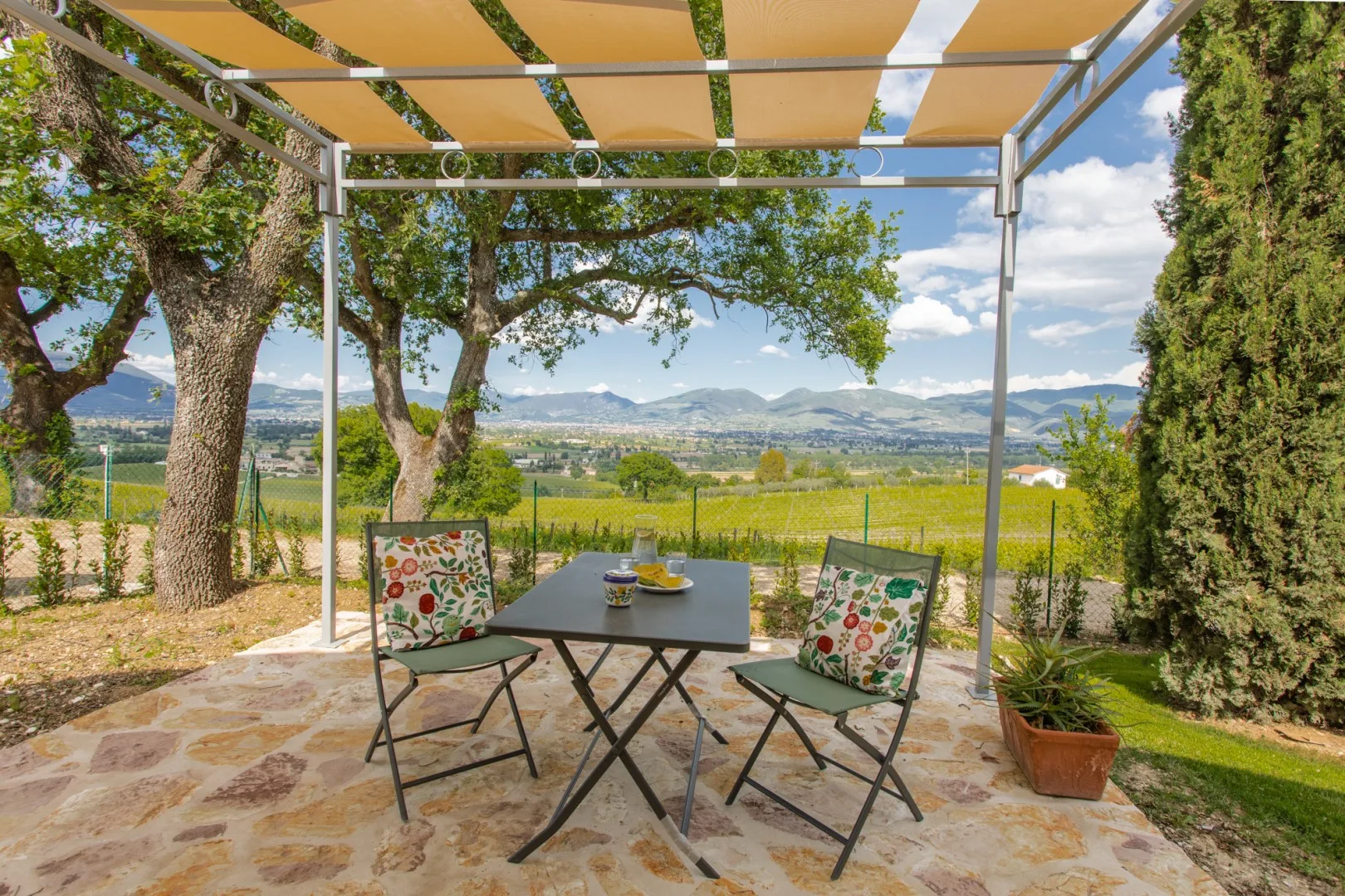 A shaded outdoor patio has a small table and seating overlooking a green valley.