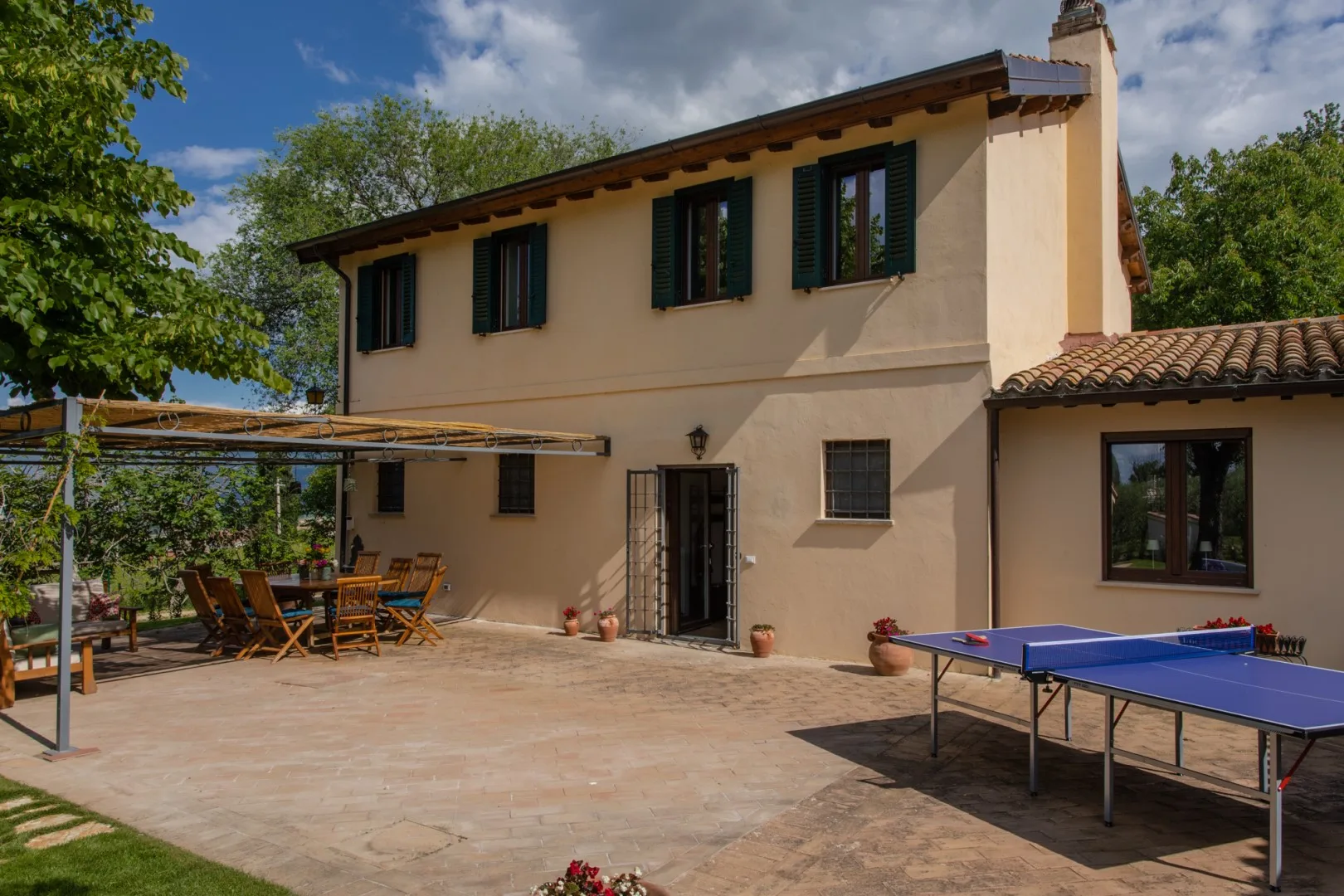 A two-story house opens onto a paved patio with outdoor seating and a table tennis table.
