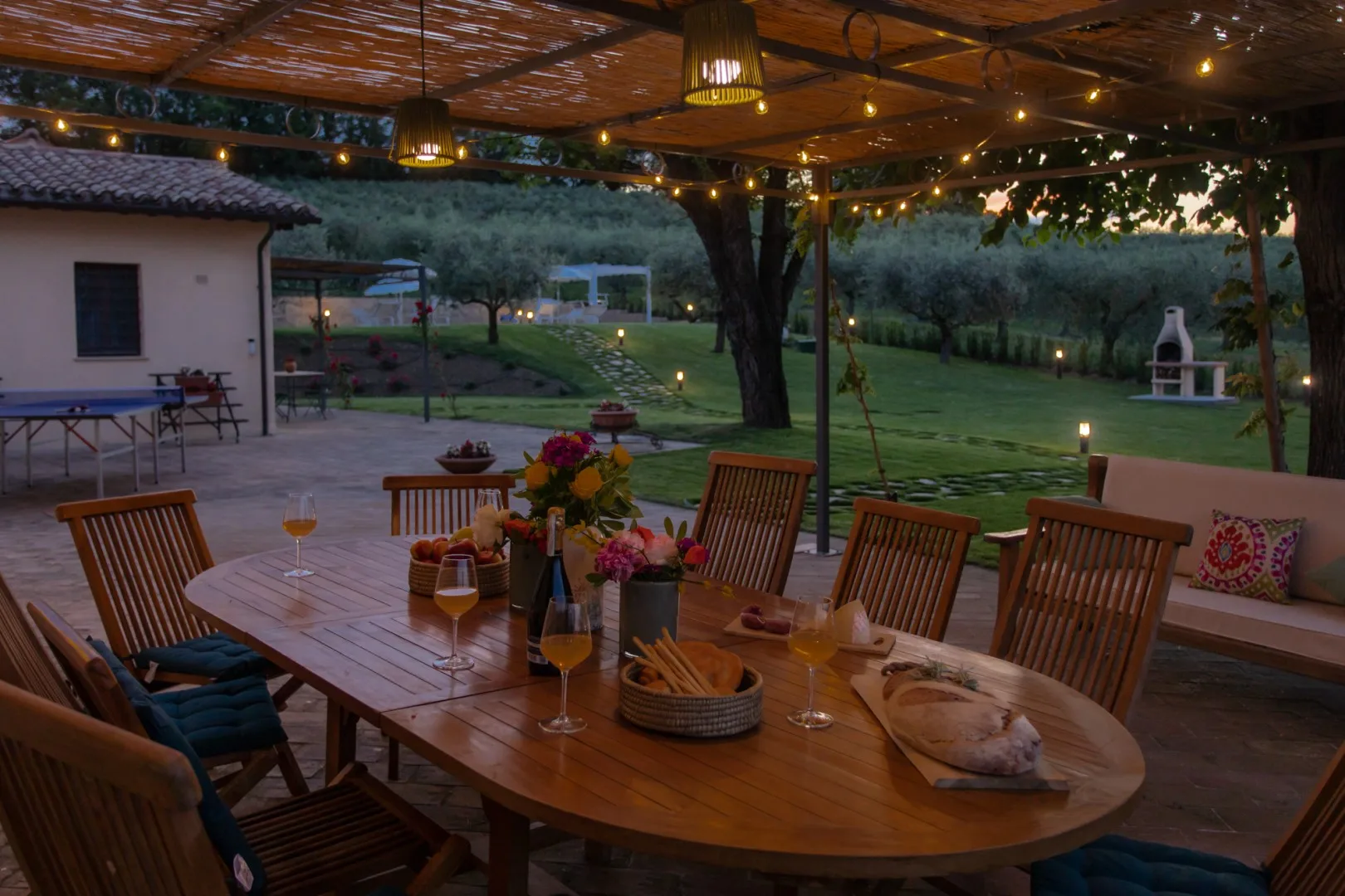 An outdoor dining area beneath a covered patio, overlooking a garden at dusk.