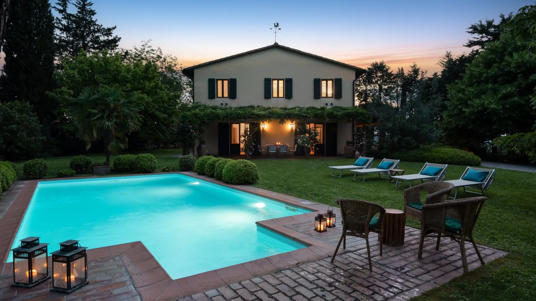 Una casa y una piscina iluminada están rodeadas de árboles al anochecer.