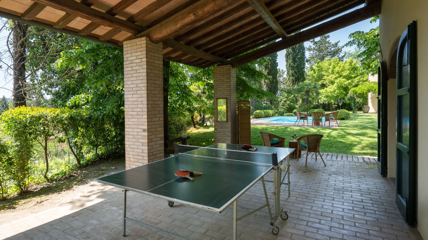 Un patio cubierto con una mesa de ping-pong frente a un jardín y una piscina.