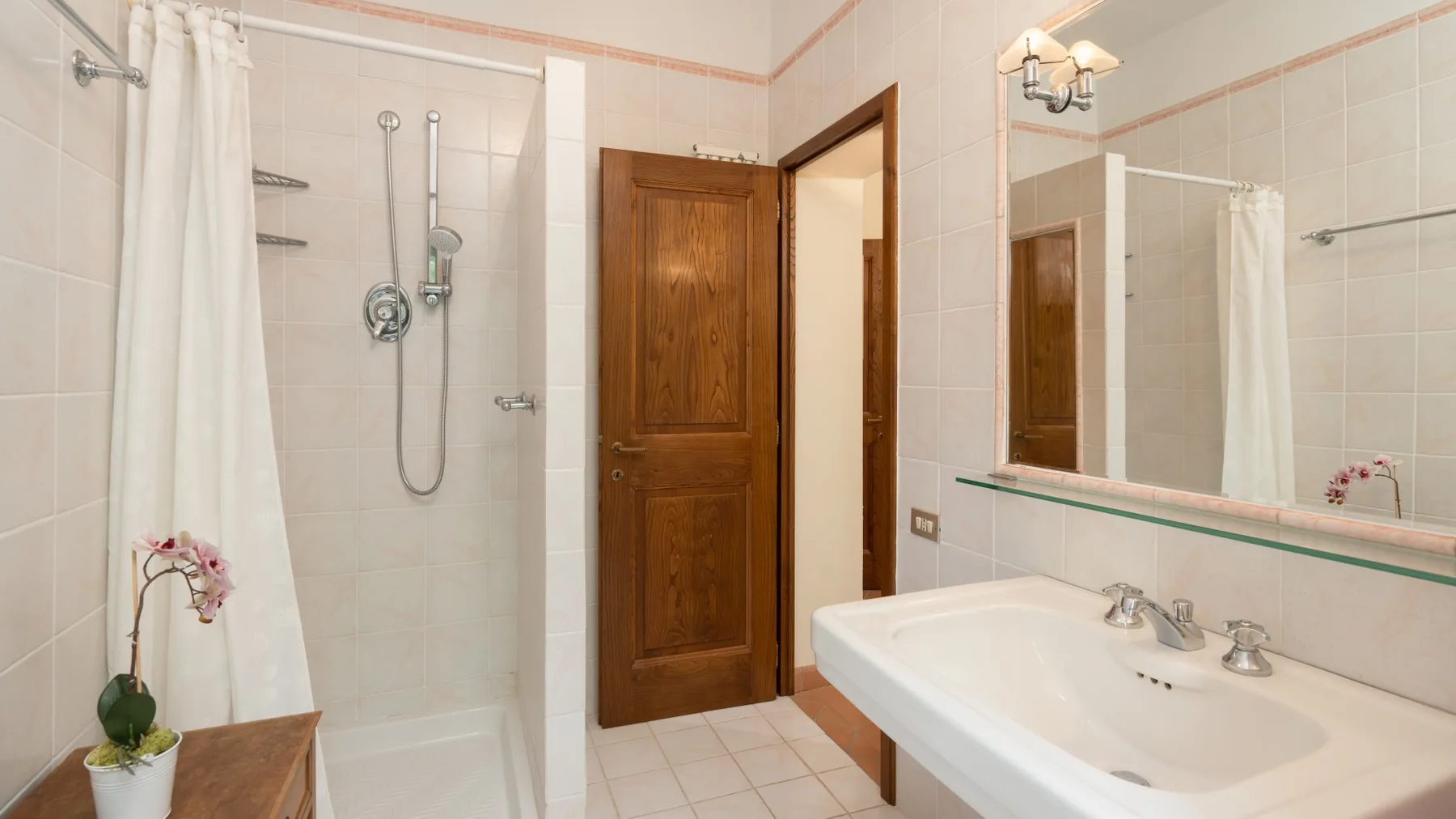 Un baño con azulejos, ducha, puerta de madera y lavabo bajo un espejo.