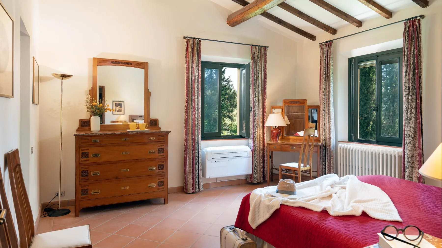 Un dormitorio con una cama cubierta de rojo, una cómoda de madera y ventanas altas.