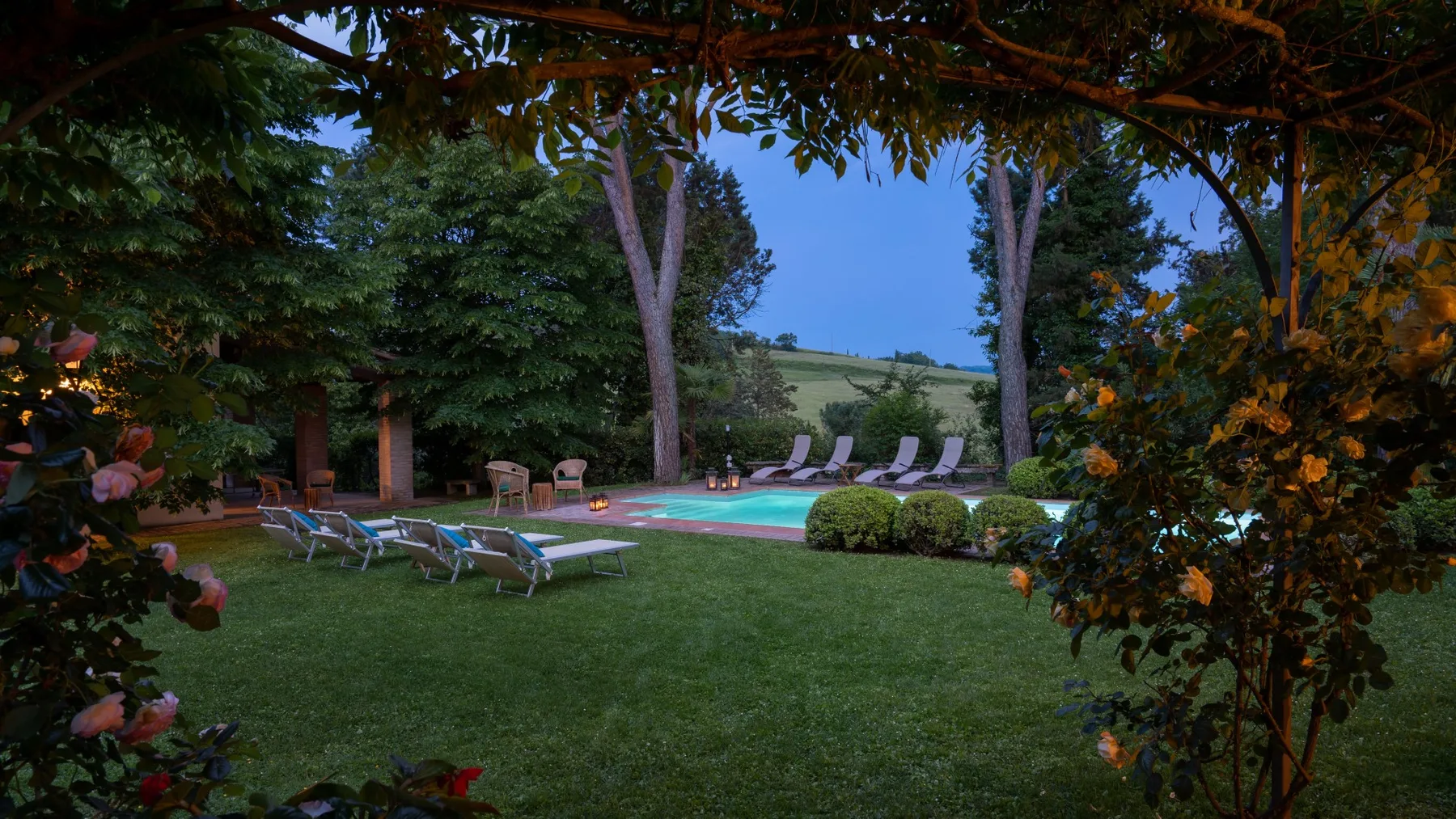 Una piscina en el jardín está rodeada de césped, asientos y árboles frondosos.
