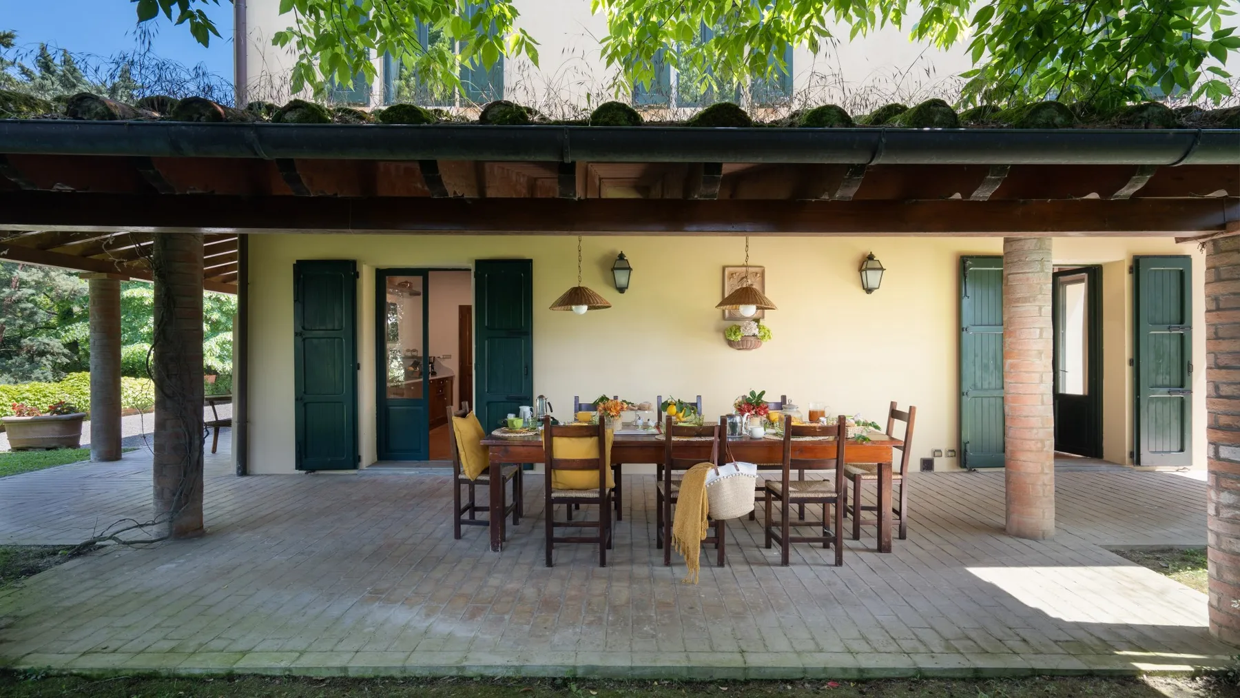 Un patio cubierto tiene una larga mesa de comedor con sillas y platos.