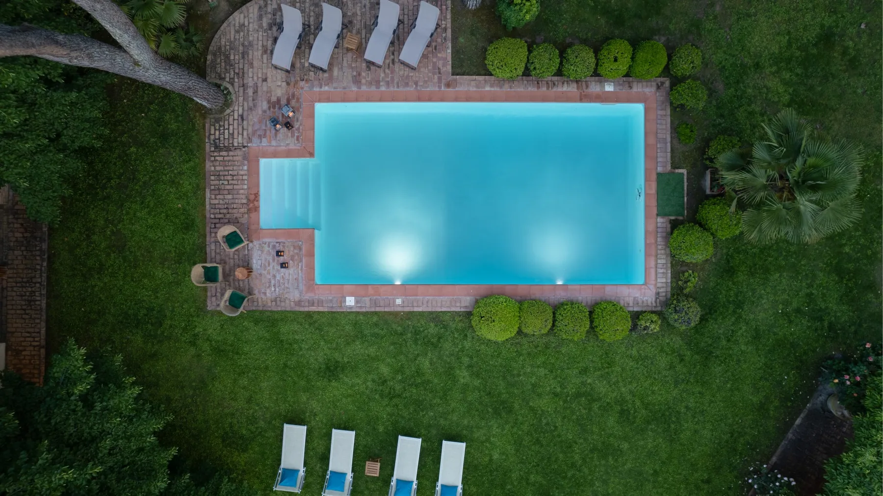 Una piscina al aire libre está rodeada por una terraza pavimentada y césped verde.