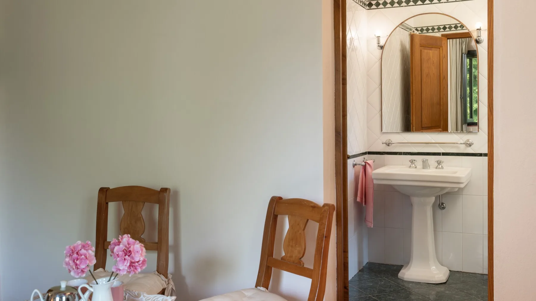Un baño con lavabo de pedestal y espejo junto a sillas de madera.