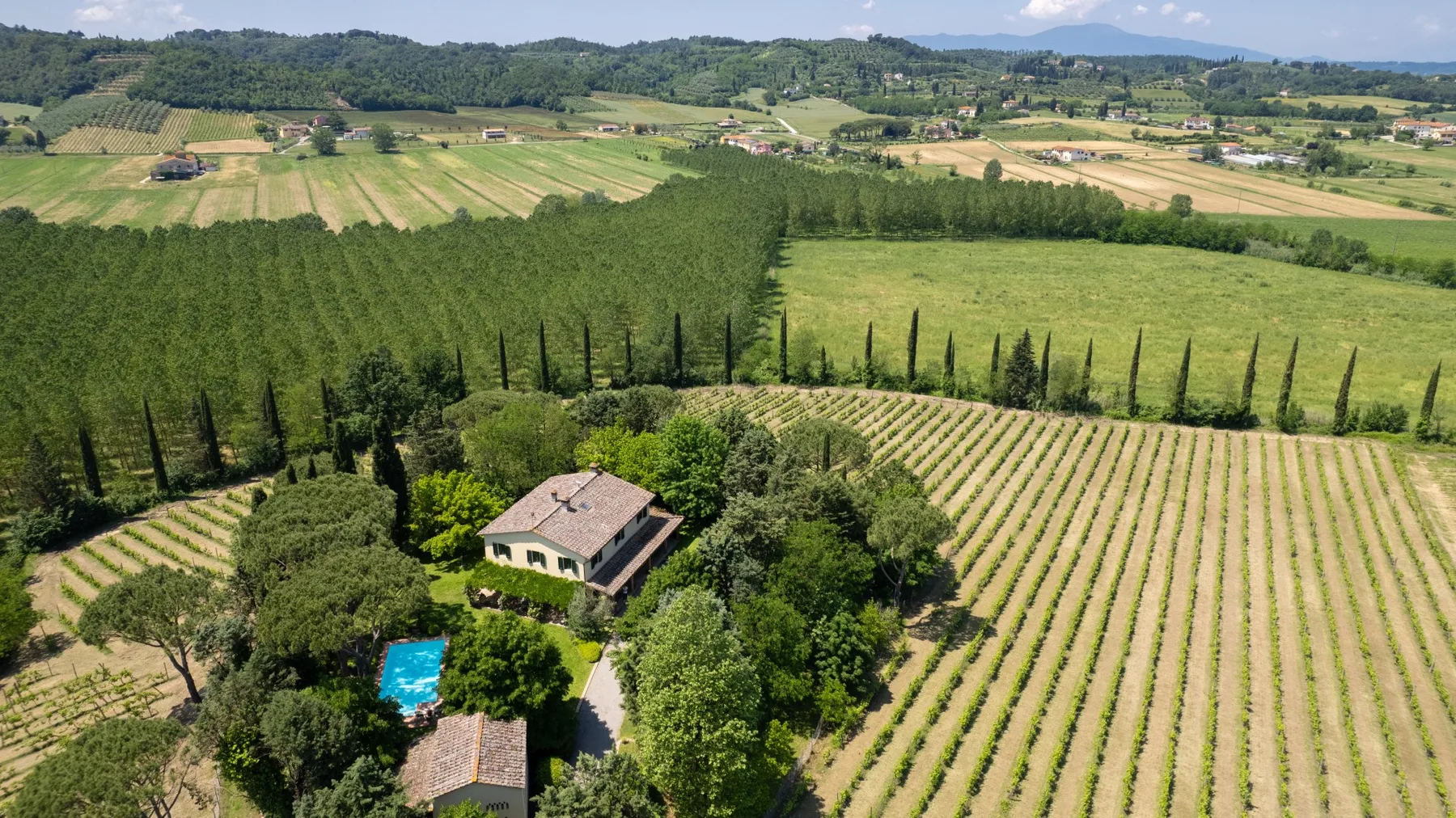 Una casa rural y una piscina azul están rodeadas de campos verdes y hileras de cultivos.