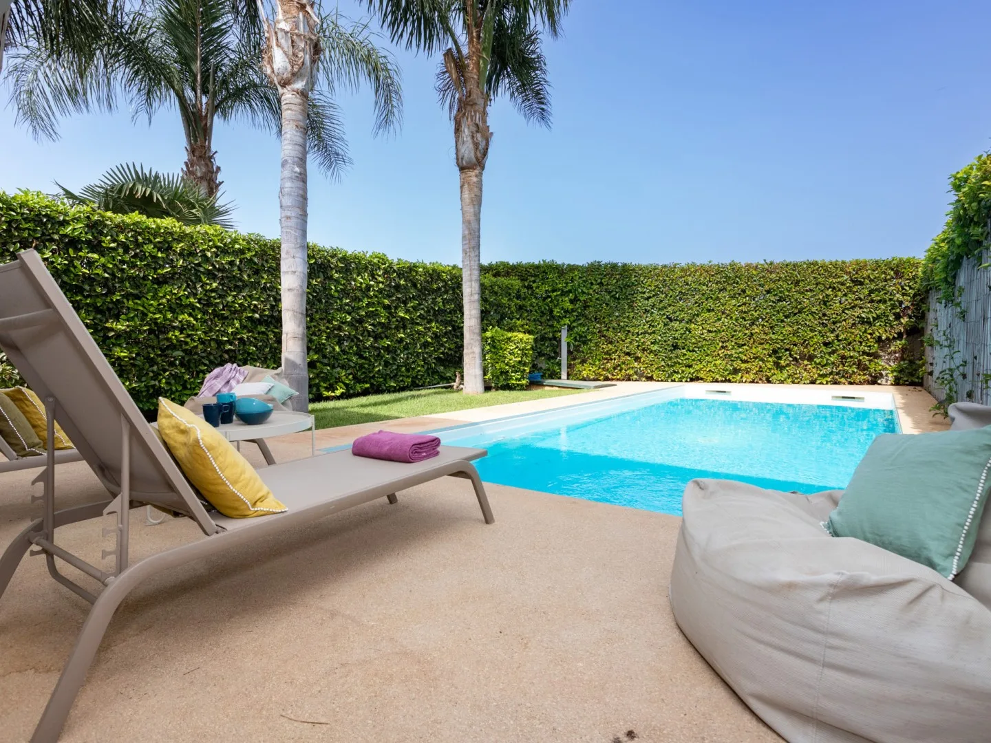 Una piscina está junto a una terraza pavimentada con tumbonas y altas palmeras.