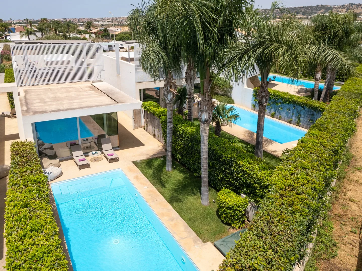 Una piscina al aire libre junto a un edificio blanco moderno, palmeras y setos recortados.