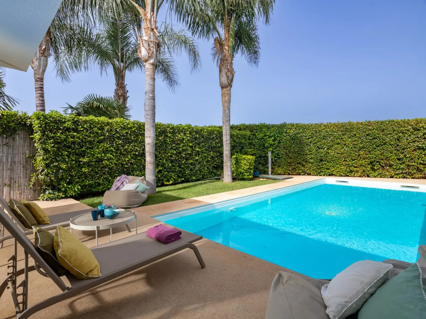 Una piscina exterior luminosa está rodeada de palmeras y asientos de patio.