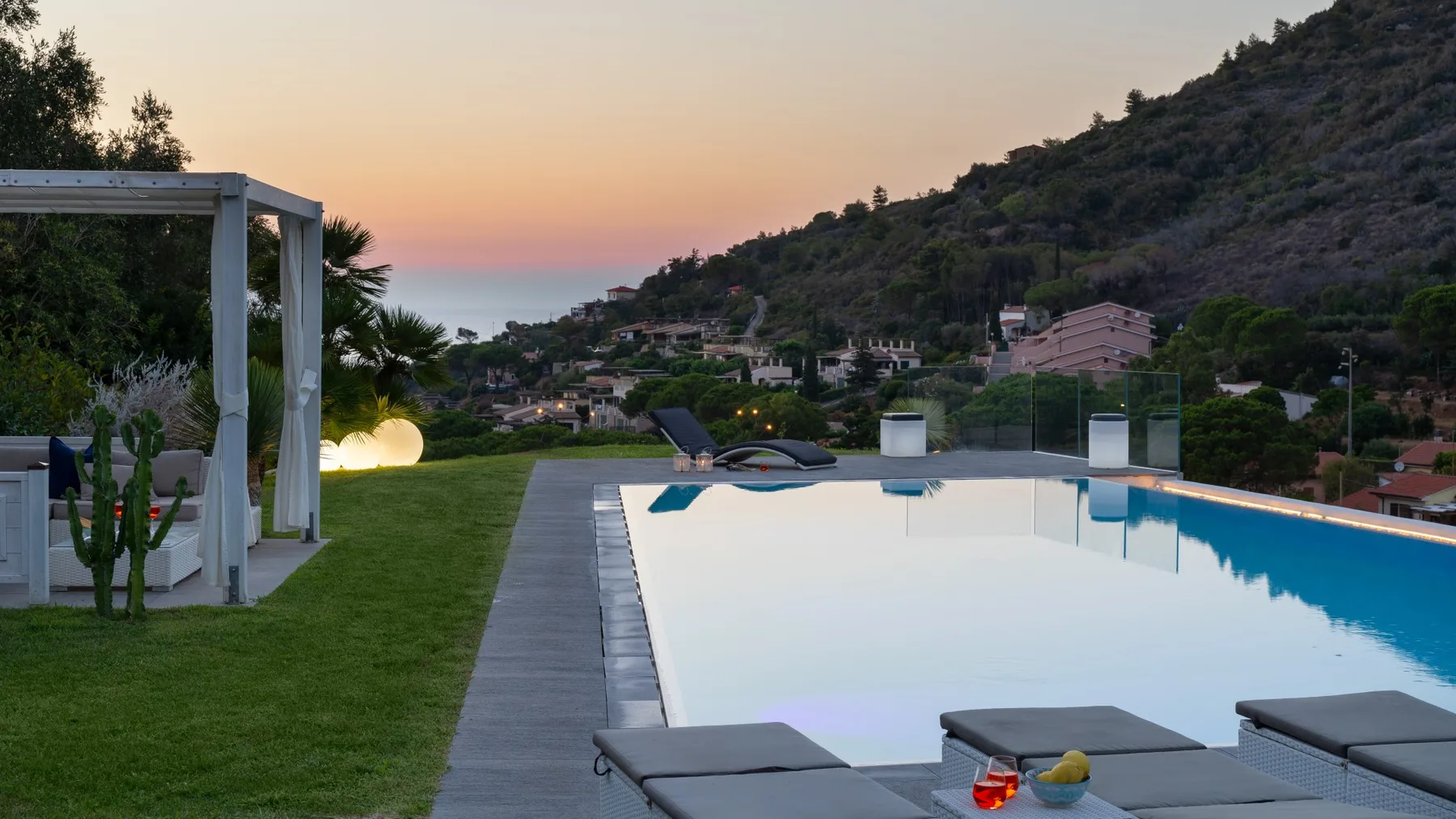 Una piscina al aire libre junto a un césped ofrece vistas de casas en la ladera al atardecer.