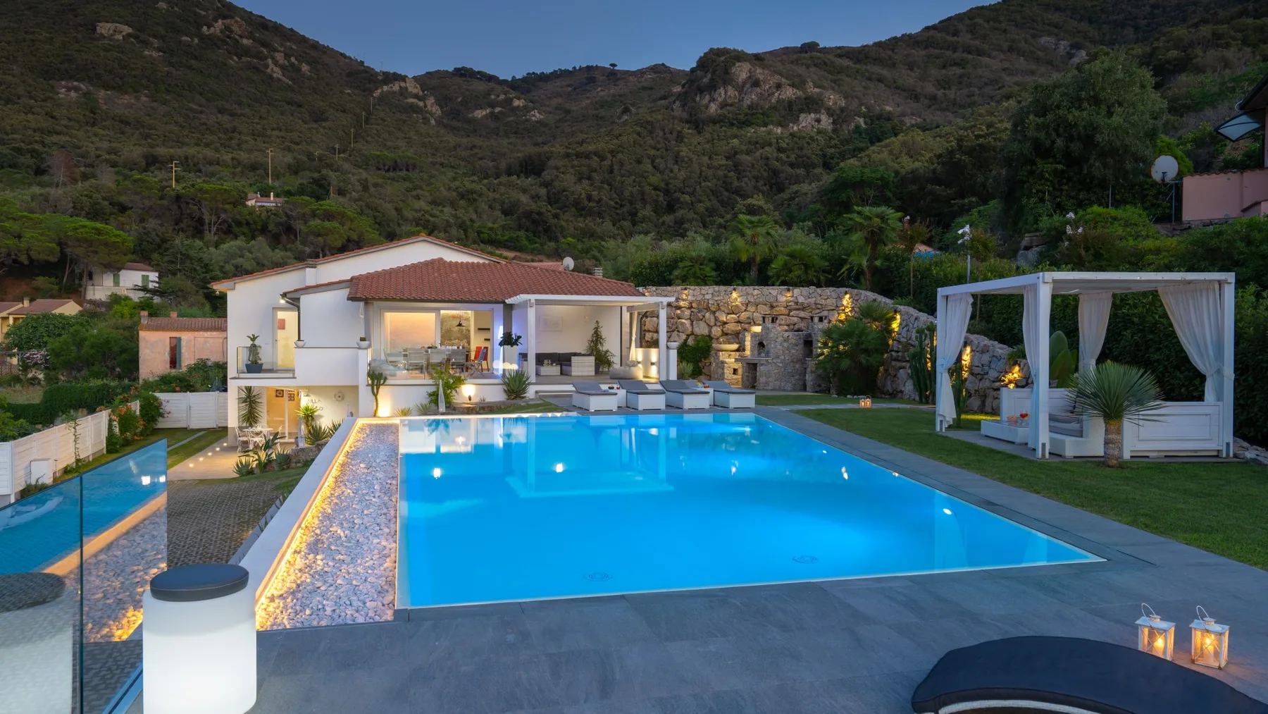 Una piscina con el borde iluminado está junto a una villa ajardinada.