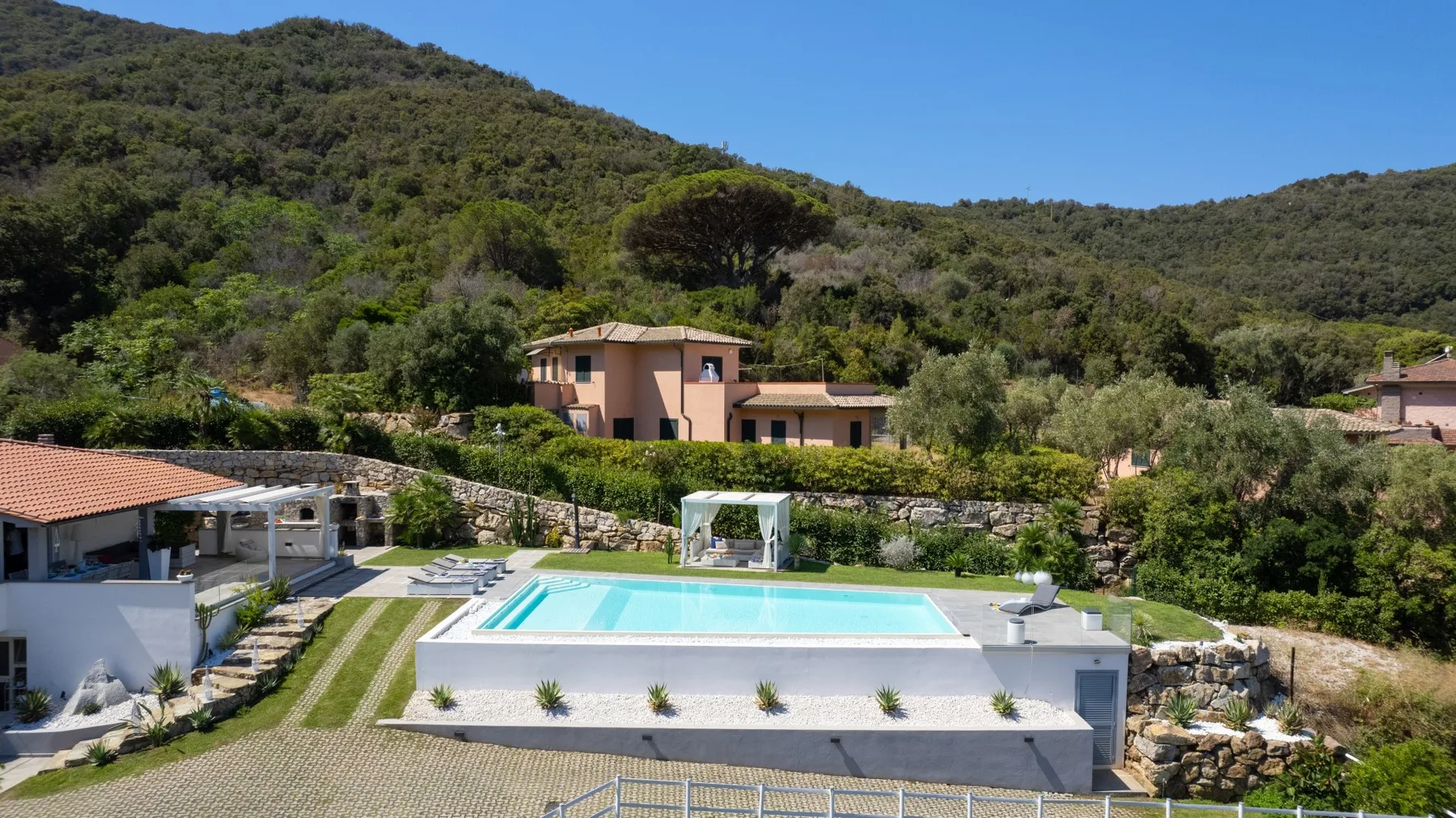 Una piscina al aire libre está frente a una casa entre colinas arboladas.