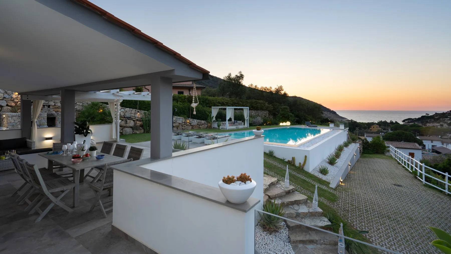 Una zona de comedor exterior cubierta da a una piscina y al atardecer.