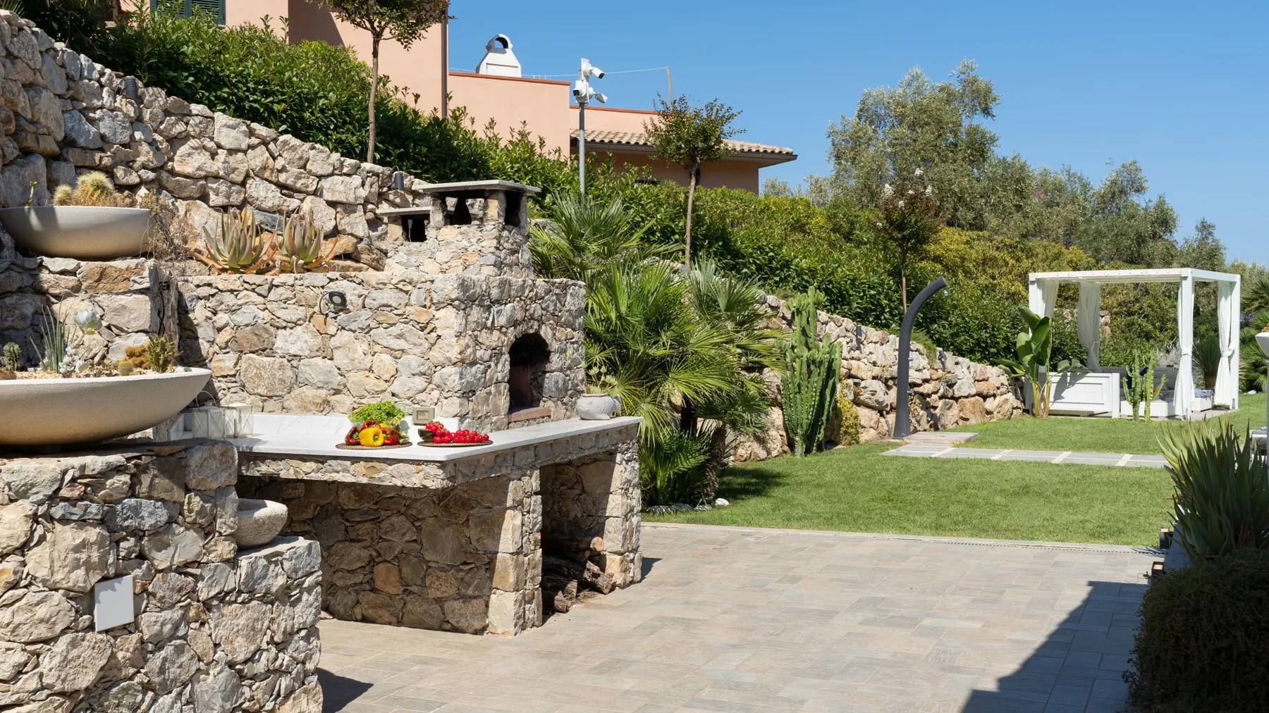 Una zona de cocina exterior de piedra da a un césped y asientos blancos.