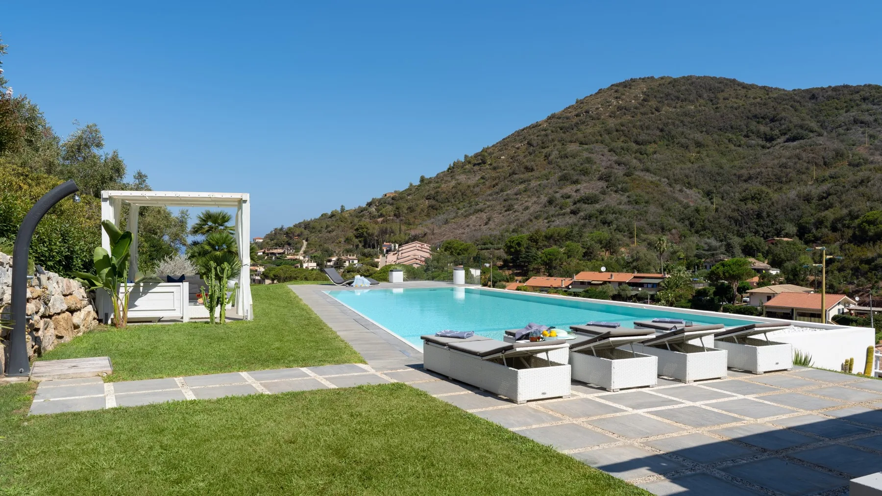 Una piscina al aire libre junto a una zona pavimentada con asientos y una colina verde al fondo.