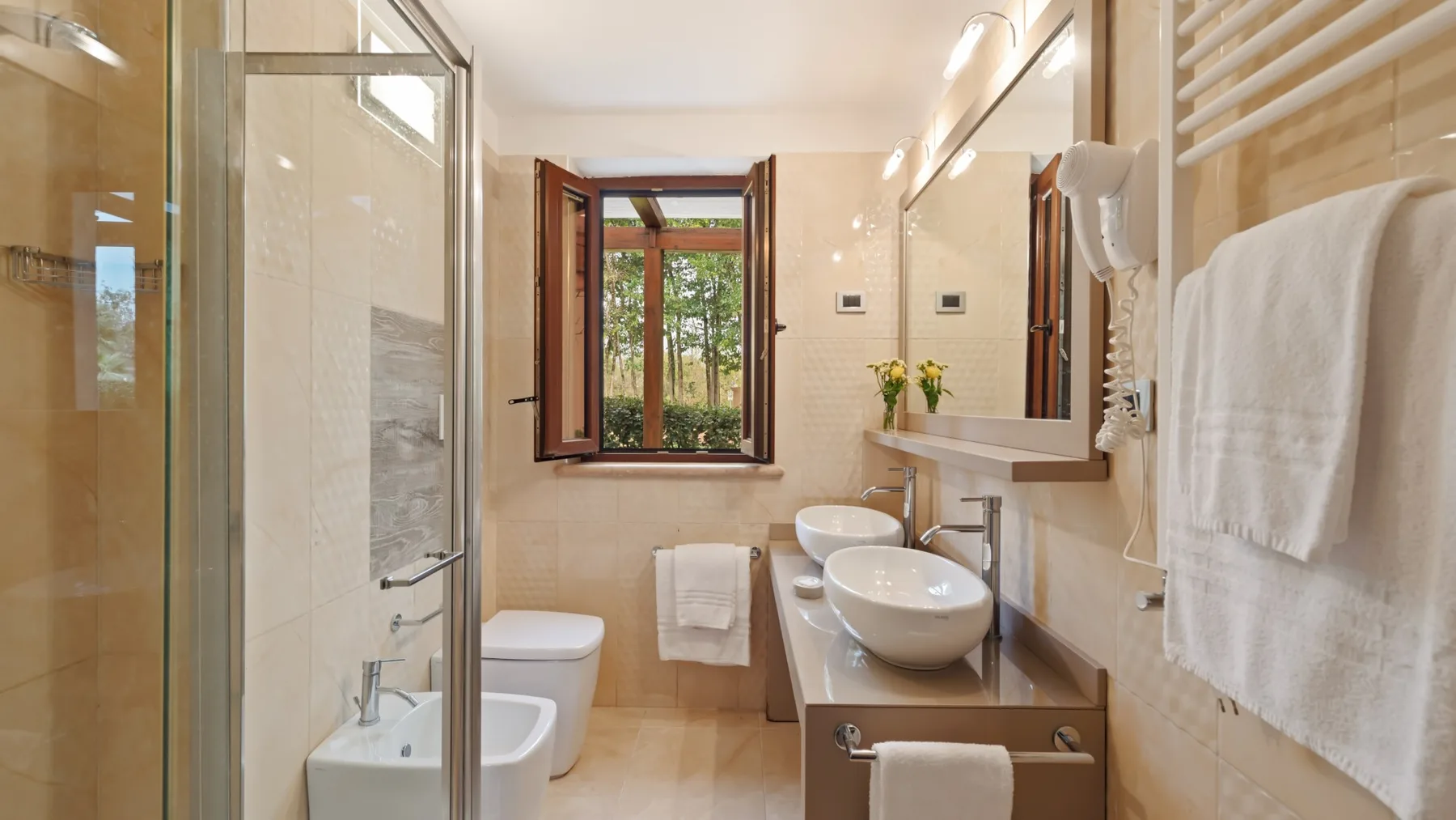 A bright bathroom with a glass shower, sinks, and a window overlooking greenery.