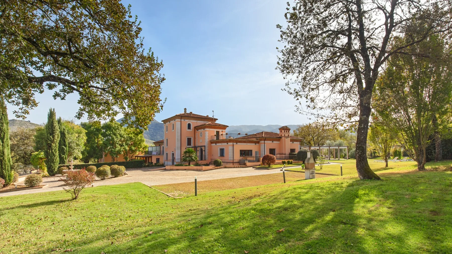 A large house stands beyond a lawn, with mature trees and hills in the distance.