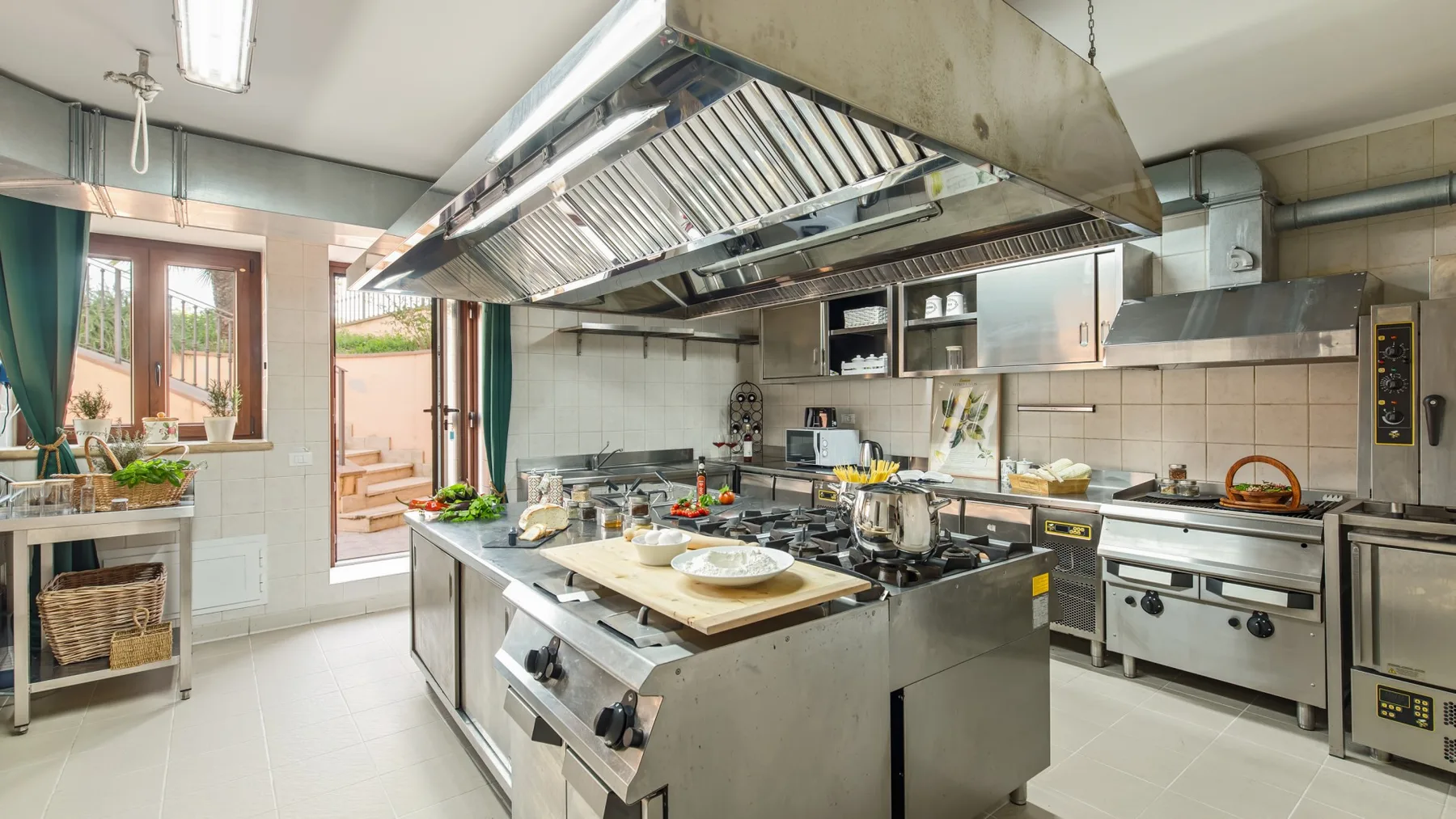 A commercial kitchen with stainless-steel counters and a large overhead ventilation hood.
