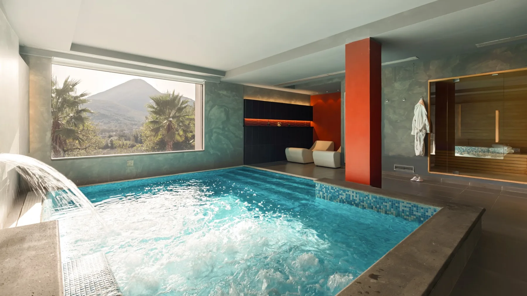 An indoor swimming pool fills a room with a large window overlooking a mountain landscape.