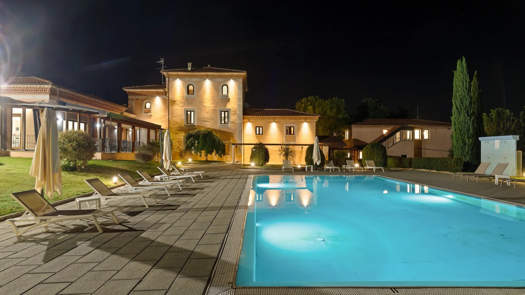 A lit building stands beside a swimming pool and paved patio at night.