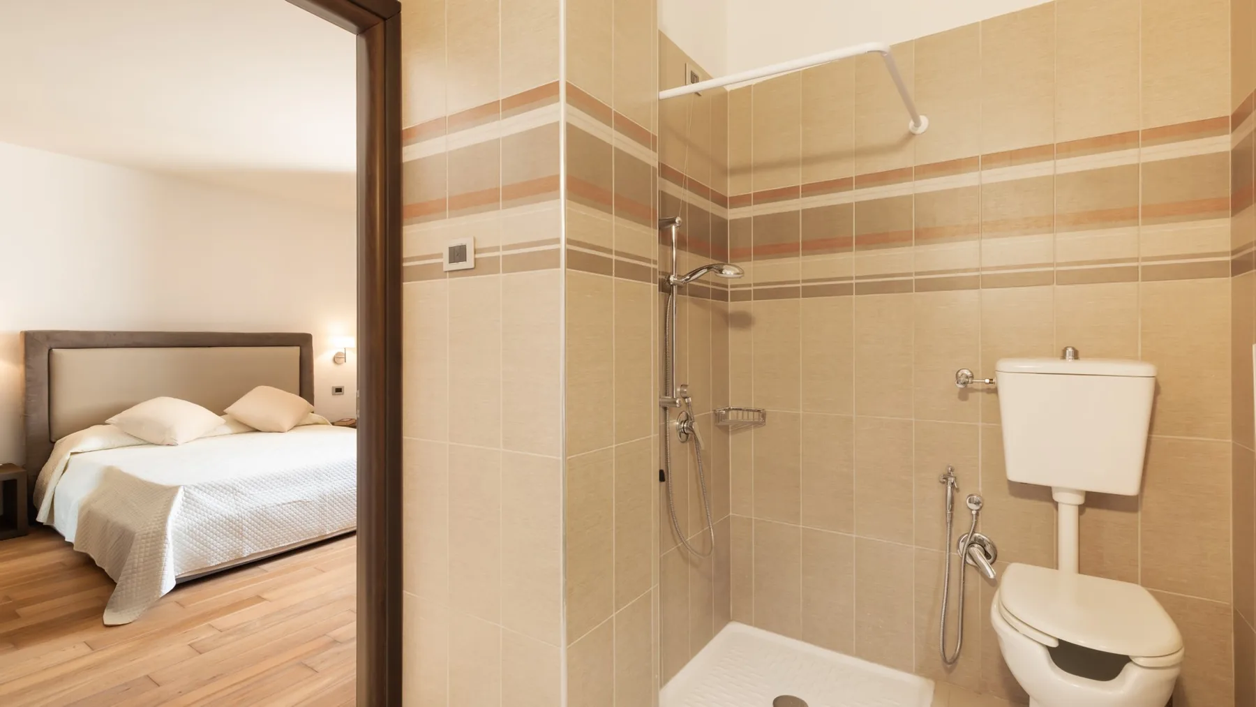 A tiled bathroom with a shower and toilet, viewed beside an adjoining bedroom.