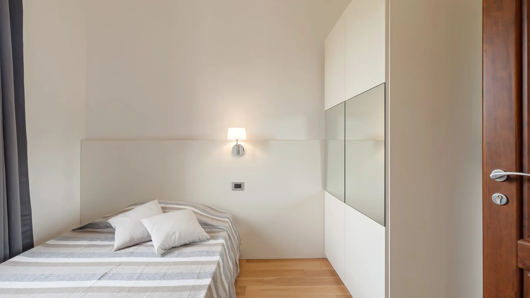 A bedroom with a bed beside a mirrored wardrobe and a wooden door.