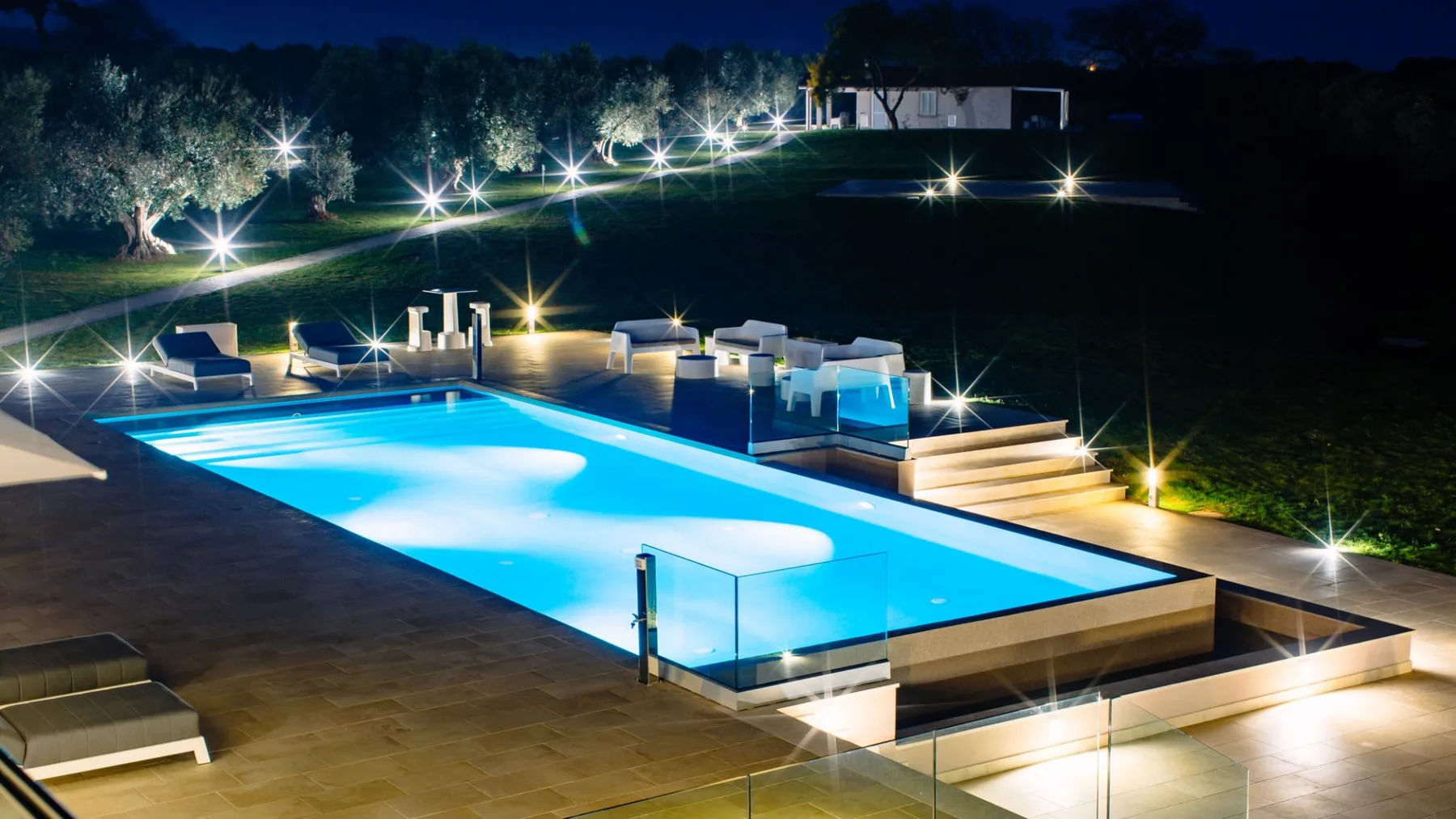Una piscina exterior iluminada con asientos y jardines durante la noche.