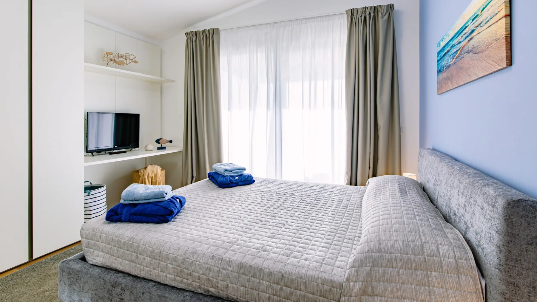 Un dormitorio con una cama grande, un televisor y cortinas frente a una ventana luminosa.