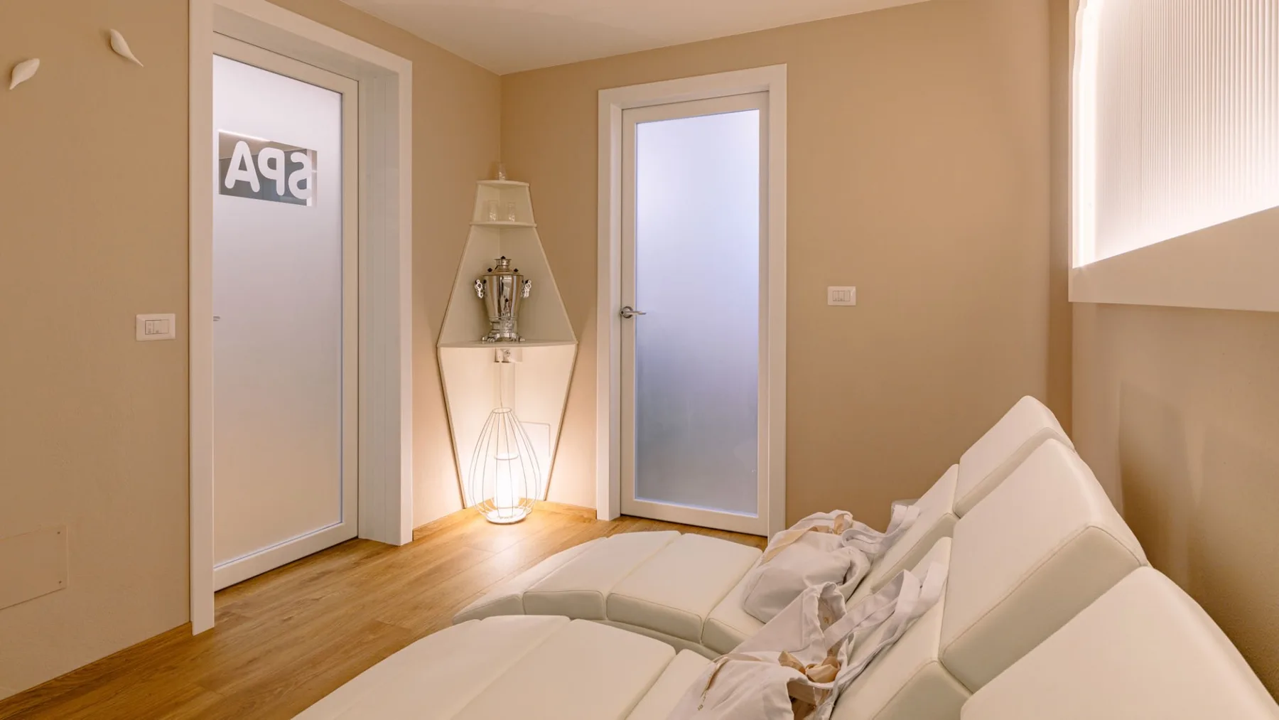 Una habitación beige con asientos blancos, dos puertas de vidrio esmerilado y una lámpara de pie.