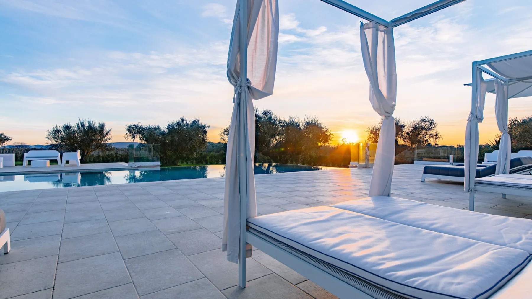 Un patio junto a la piscina con tumbonas acolchadas y el atardecer al fondo.