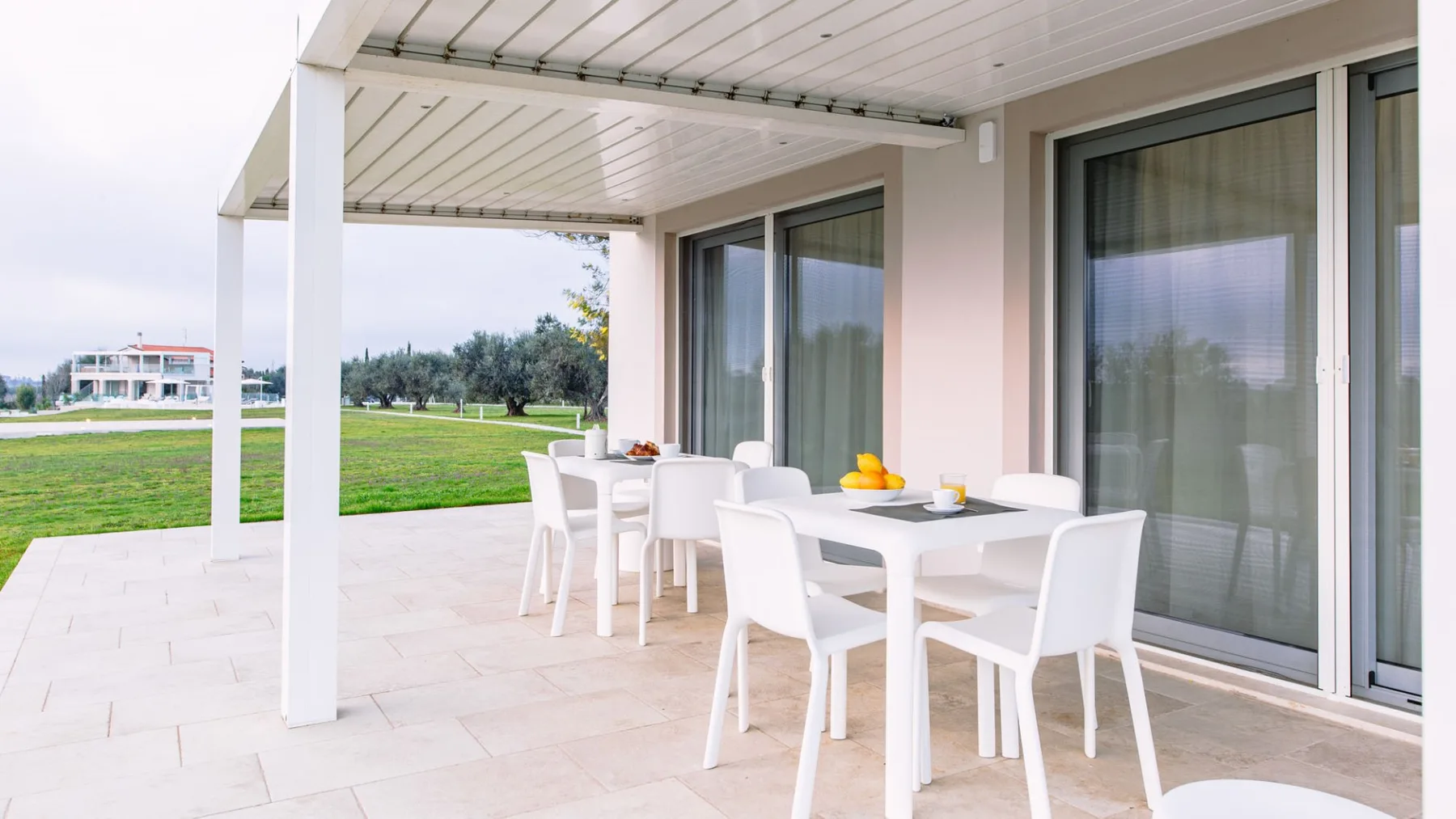 Una zona de comedor exterior cubierta con mesas y asientos blancos da a un césped verde.