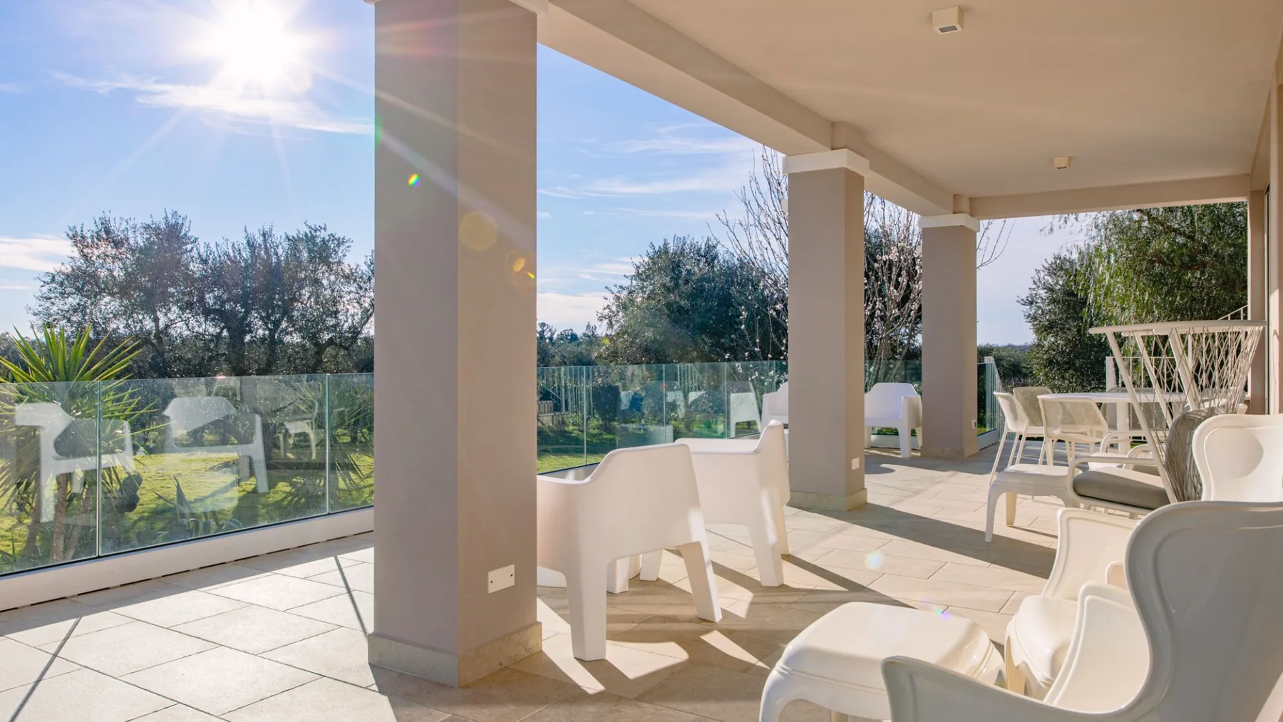 Un patio exterior cubierto con asientos blancos y una barandilla de vidrio da a un jardín.