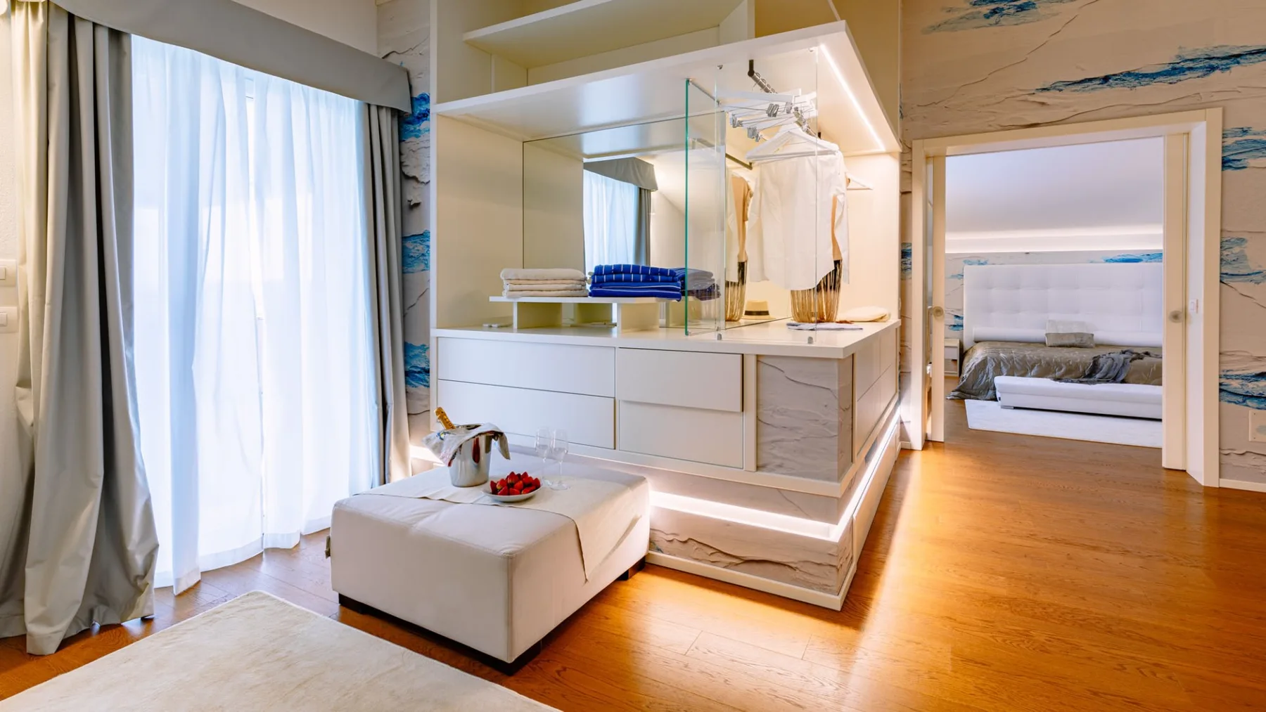 Un dormitorio con zona de vestidor, una cama visible a través de la puerta y una gran ventana con cortinas.