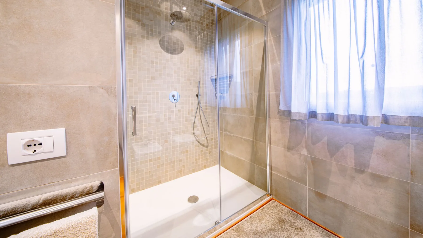 Un baño con azulejos, una ducha con mampara de vidrio y una ventana con cortinas.