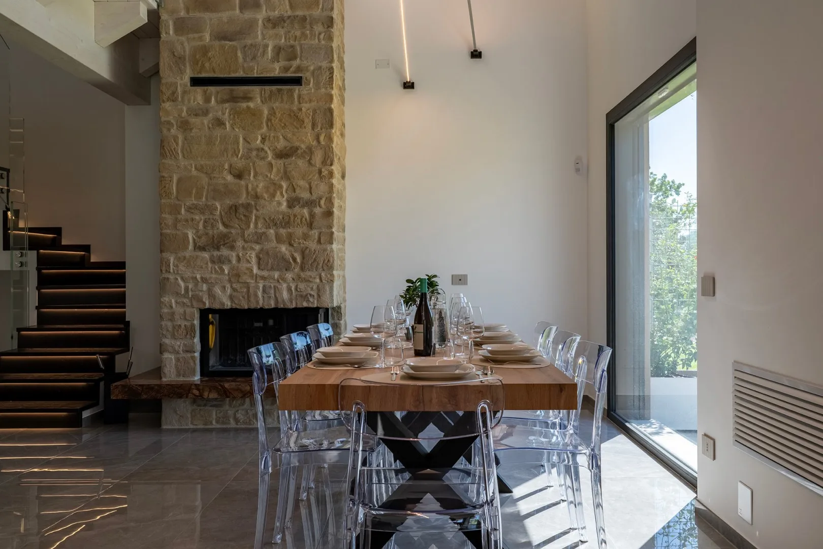 Una larga mesa de comedor está puesta con platos y copas junto a una chimenea de piedra y una puerta de vidrio.