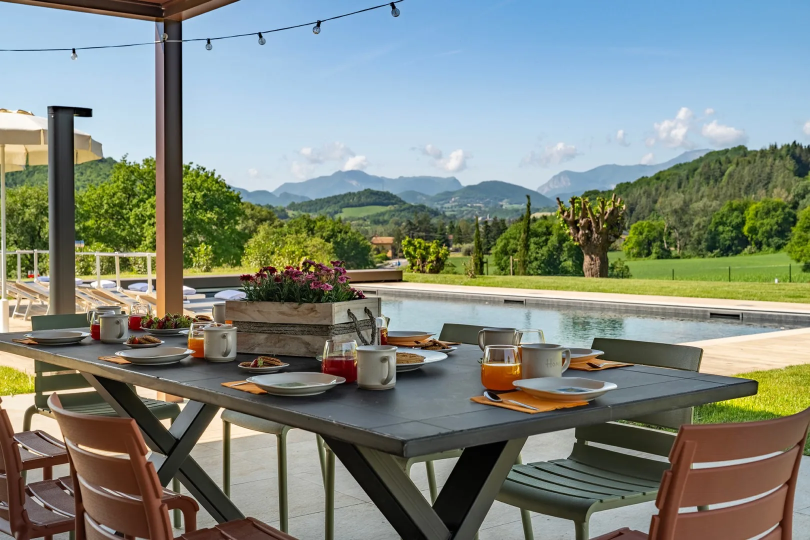 Una mesa de comedor al aire libre está junto a una piscina, con colinas verdes a lo lejos.