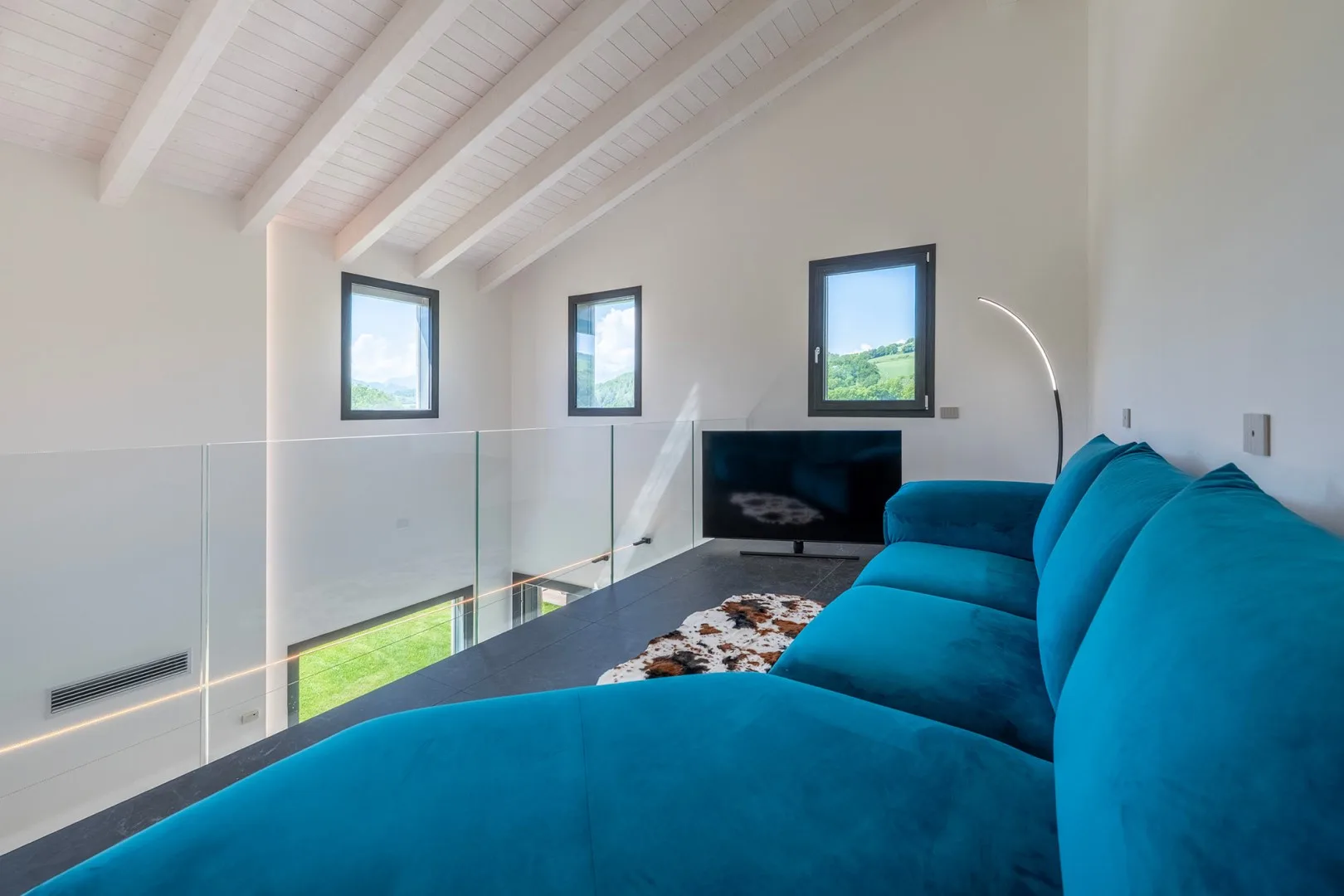 Una luminosa sala tipo loft tiene un gran sofá seccional turquesa, un televisor y ventanas bajo un techo blanco inclinado.
