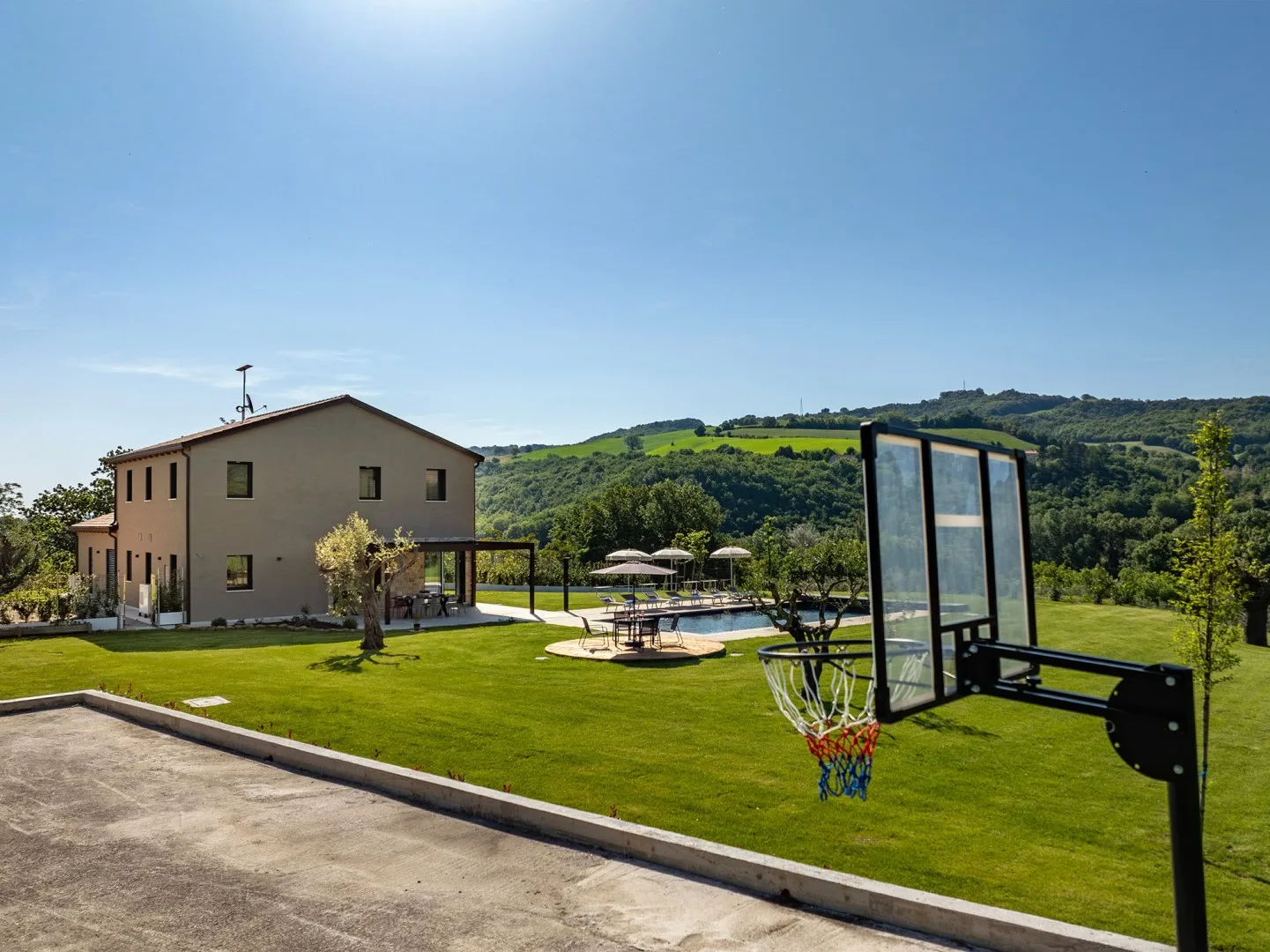 Una casa moderna da a un césped verde con una canasta de baloncesto, asientos al aire libre y colinas.