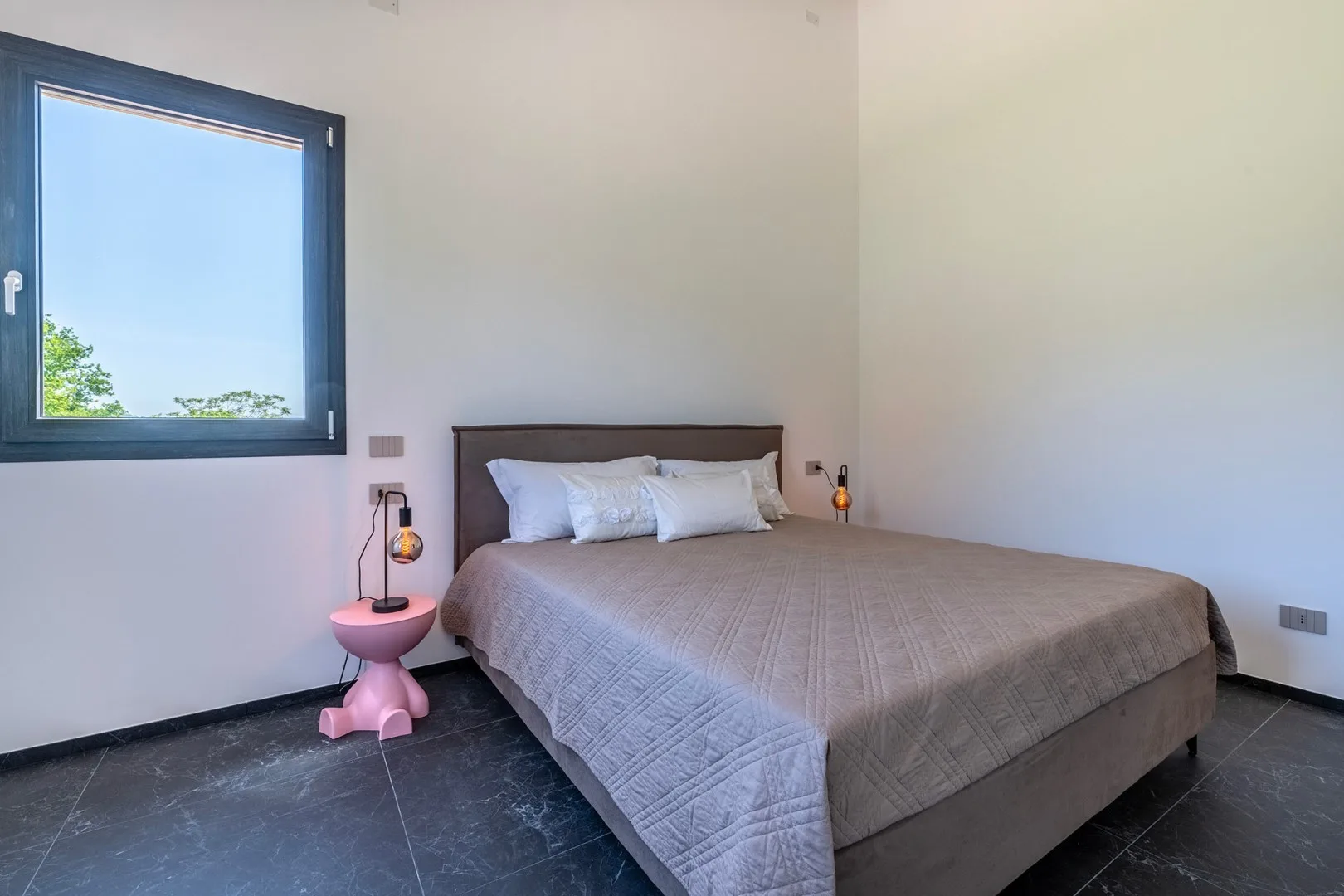 Un dormitorio con una cama doble, colcha gris, almohadas blancas y una ventana con vistas a la vegetación.
