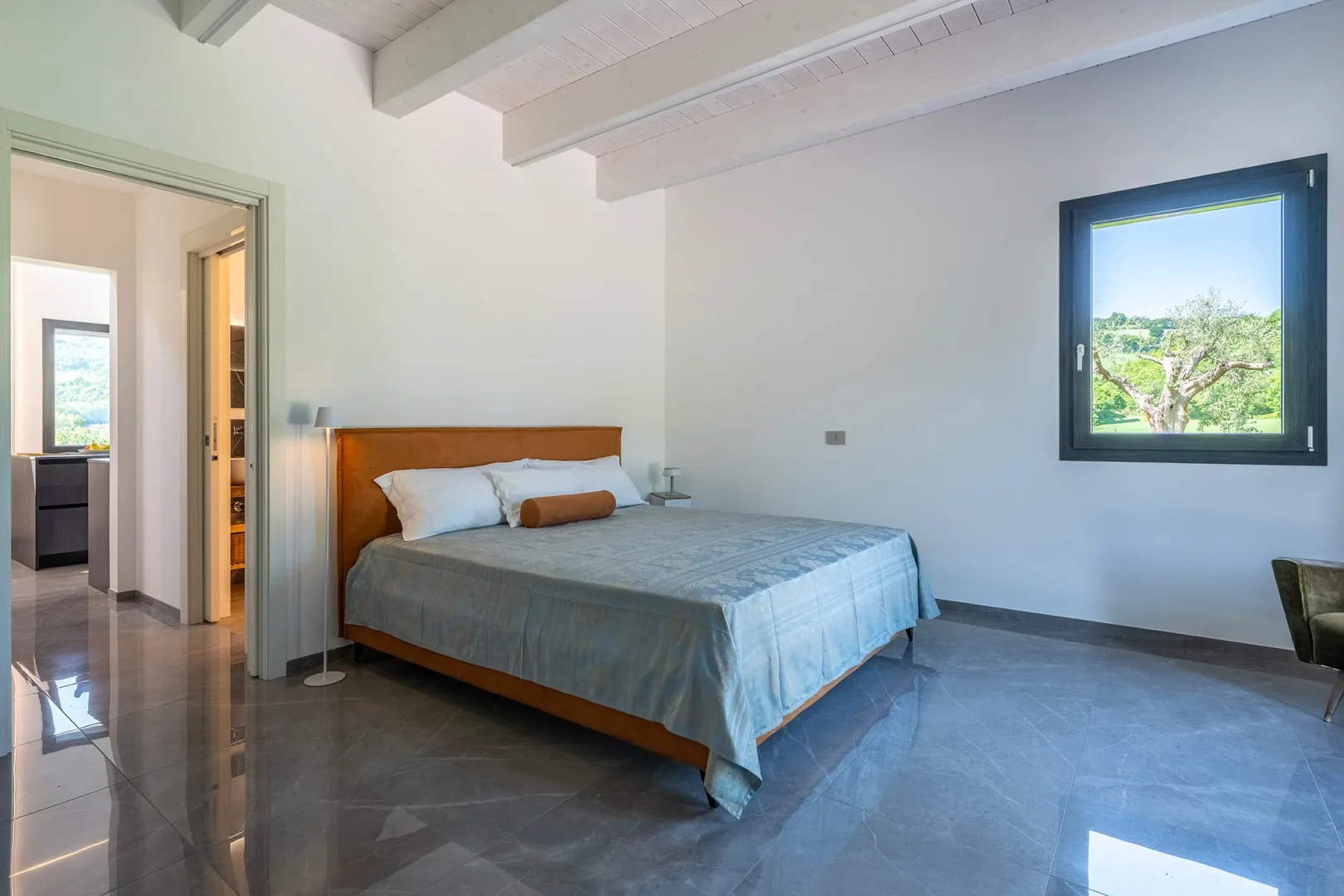 Un dormitorio con una cama de madera, ropa de cama gris, vigas blancas en el techo y una ventana con vistas a la vegetación.