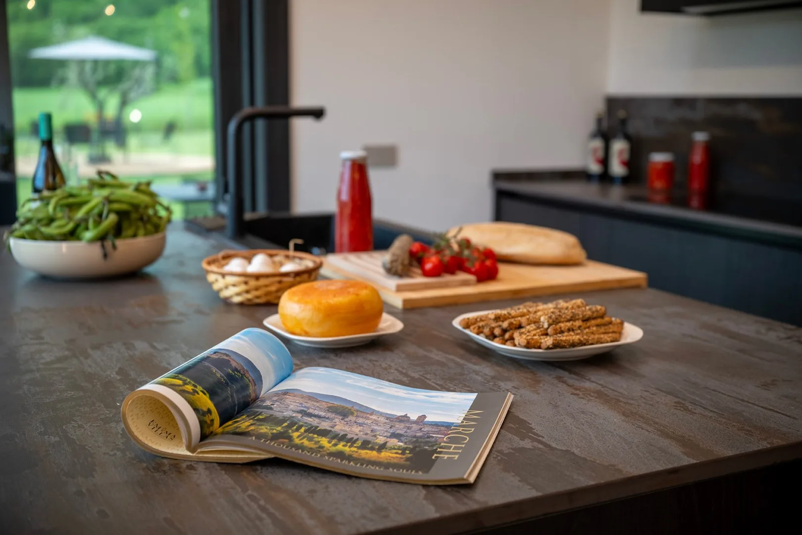 Una isla de cocina muestra productos frescos, pan, salchichas y una revista de viajes abierta junto a una gran ventana.