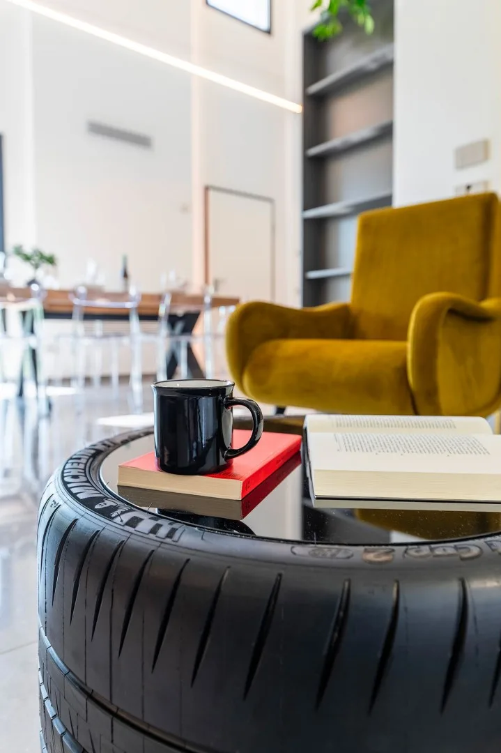 Una mesa de centro con forma de neumático sostiene una taza y libros junto a un sillón amarillo mostaza en un comedor luminoso.
