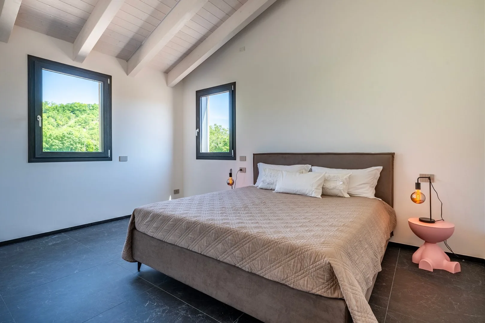 Un dormitorio con una cama grande, dos ventanas y vigas blancas a la vista en el techo.