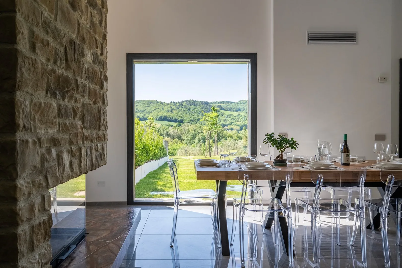 Una mesa de comedor con sillas transparentes está junto a un gran ventanal con vistas a colinas verdes y un muro de piedra.