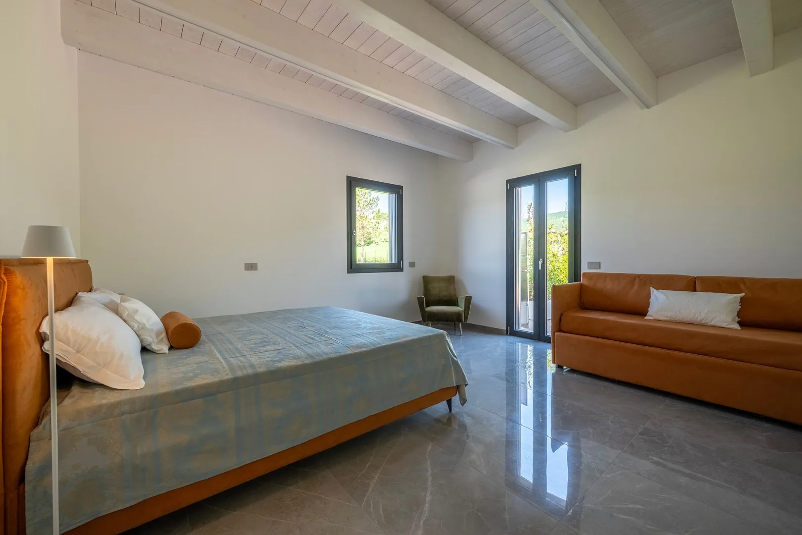 Dormitorio con una cama doble, un sofá marrón y puertas acristaladas altas con vistas al exterior.